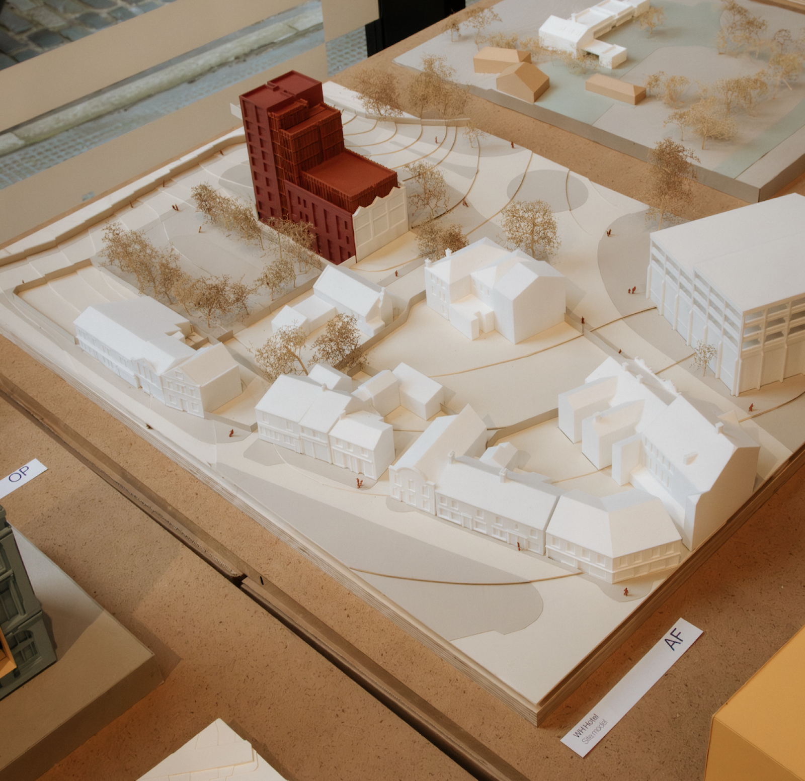 Wolverhampton House. Scale 1:200. Site Model

As a site model, this was useful in communicating the impact of the proposal on its surrounding buildings, particularly given its location next to a Grade II listed building. The model was used within the Design and Access Statement and for social media, including the creation of a reel that documented the model-making process.

Due to the contrast between the white PLA 3D-printed existing elements on site and the spray painted areas of the new build, the proposal is distinguished from its context and clearly highlights the retention of the existing façade and the introduction of new construction behind it. The variation in the colour of the spray paint also communicates material differences, with the darker red indicating the brick volume and the orange representing metal-clad elements.

Plywood, PLA 3D Print, mountboard, sea foam trees, spray paint, plastic people

Made by Camila Alvarado, Part 2 Architectural assistant 
Eve Harvey, Part 1 Architectural assistant 